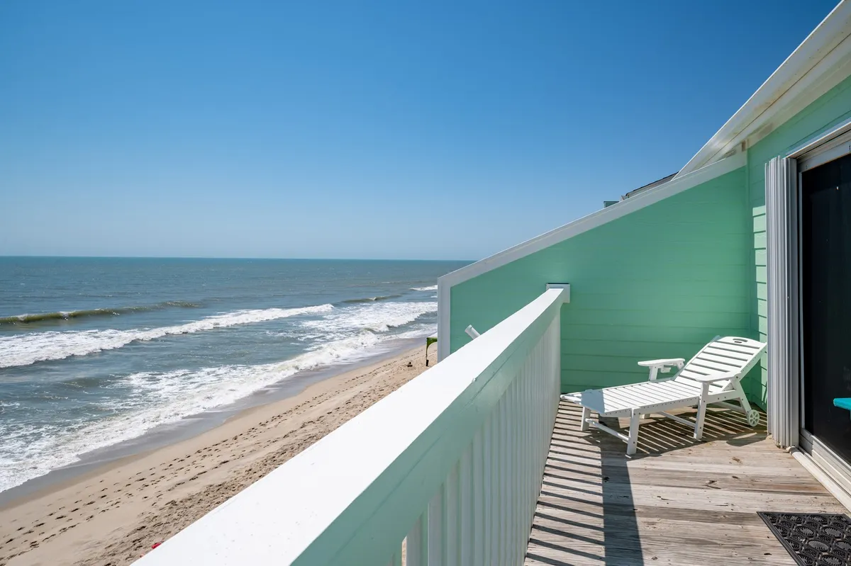 A tranquil exterior shot showcasing the expansive sandy beach and ocean, with a lounge chair and deck railing in the foreground. The soft light suggests either sunrise or early evening, and the beach appears inviting for families to play, swim, or spot wildlife.
