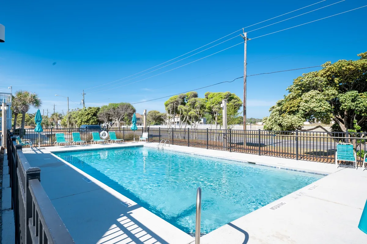 A relaxed exterior scene featuring a sparkling swimming pool surrounded by lounge chairs, umbrellas, and lush greenery. The atmosphere promotes sunbathing, swimming, and socializing in a resort-like setting.