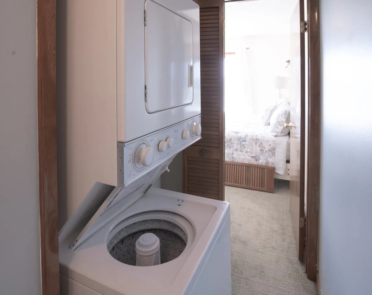 A compact laundry area featuring a front-loading washing machine and dryer, with a view into an adjacent bedroom through an open doorway. The room is organized and practical, clearly designed for easy, in-unit laundry access.