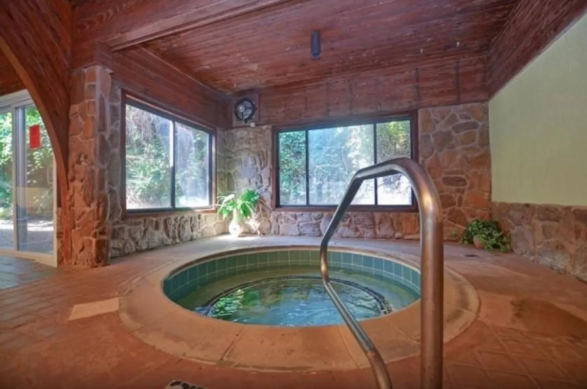 A serene indoor hot tub space features natural stone walls, large windows framing outdoor views, and lush indoor plants, creating a spa-like environment perfect for relaxation and rejuvenation.