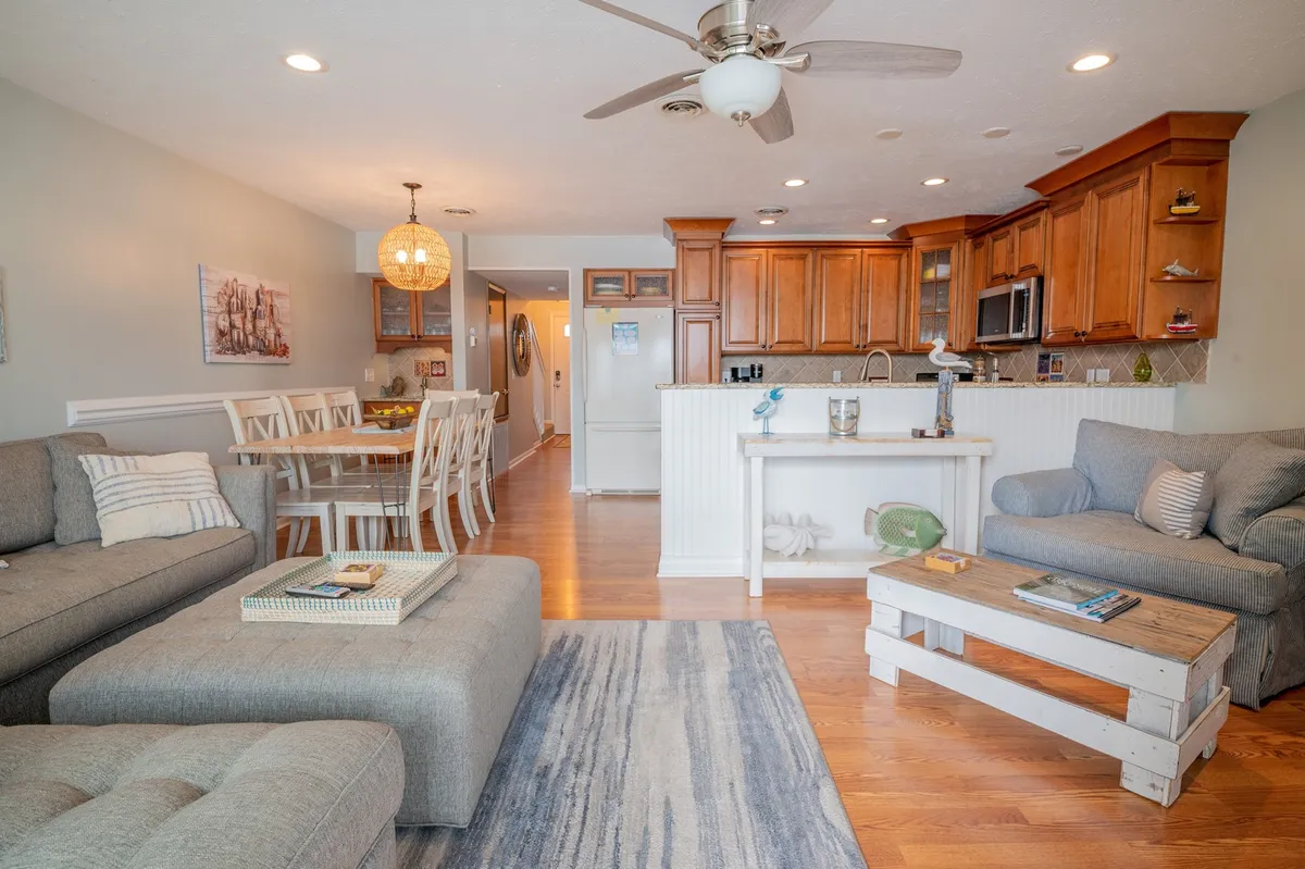 A spacious, open-concept living area featuring a comfortable sectional sofa, a large dining table with seating for multiple guests, and a kitchen island. Sunlight streams through expansive windows, creating a warm, inviting atmosphere that highlights the seamless flow between the kitchen, dining, and living spaces.