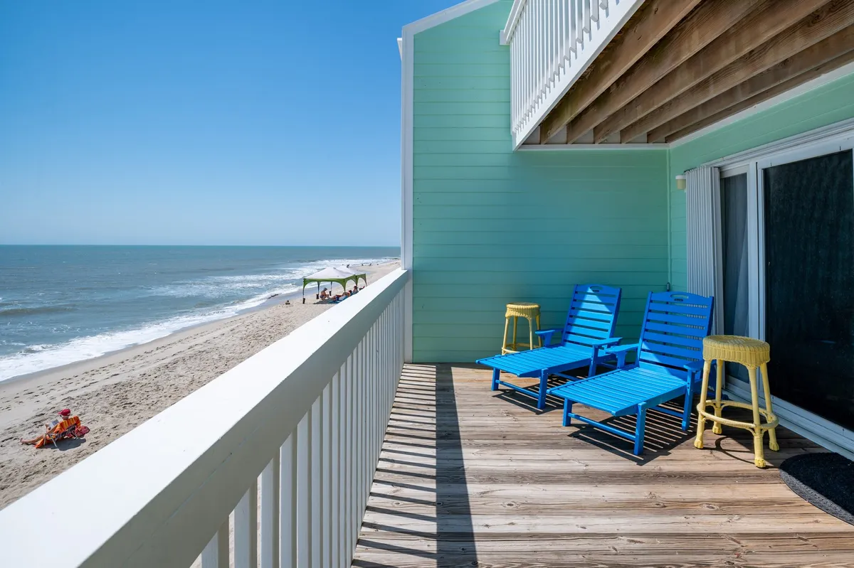 The image features a stunning oceanfront view from the condo's private deck, showcasing two inviting blue lounge chairs and a cheerful yellow accent stool. The deck overlooks the sandy beach and rolling waves, capturing the warmth of a sunny North Carolina day and inviting scenes of guests relaxing, socializing, or simply soaking up the serene coastal atmosphere.