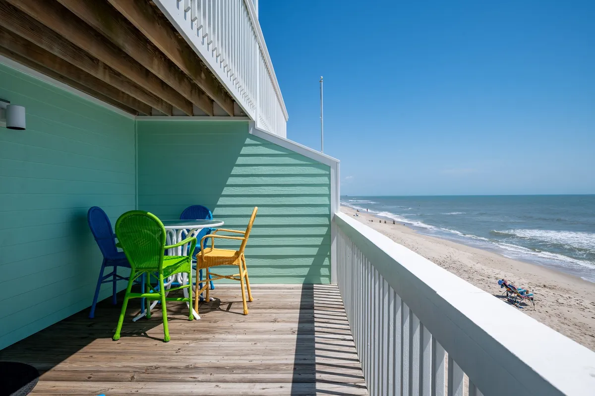 A cheerful oceanfront deck with colorful chairs and a table, overlooking the beach. The scene suggests a welcoming space for outdoor dining, relaxing, and enjoying panoramic ocean views.