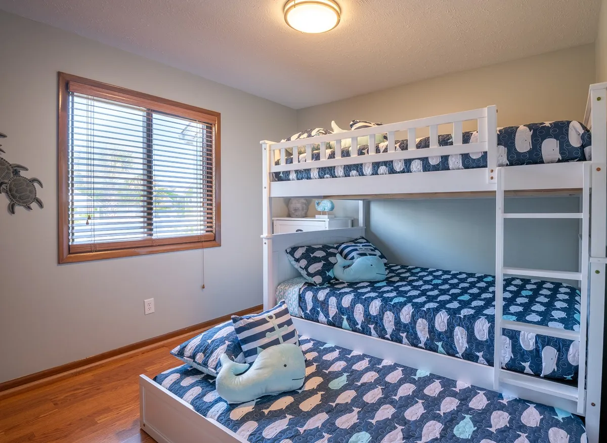 A cheerful bunk room with playful whale-themed bedding on sturdy, built-in bunk beds. A window with blinds lets in soft light, and the room is decorated to appeal to children or the young at heart, fostering a sense of fun and comfort. The setting is practical for families, offering both play and rest spaces.