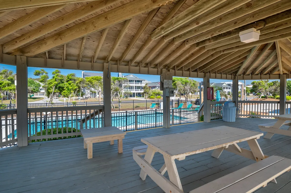 An open pavilion with wooden picnic tables, set adjacent to a pool and surrounded by greenery. The space suggests a casual, communal environment perfect for group gatherings, meals, and outdoor activities.