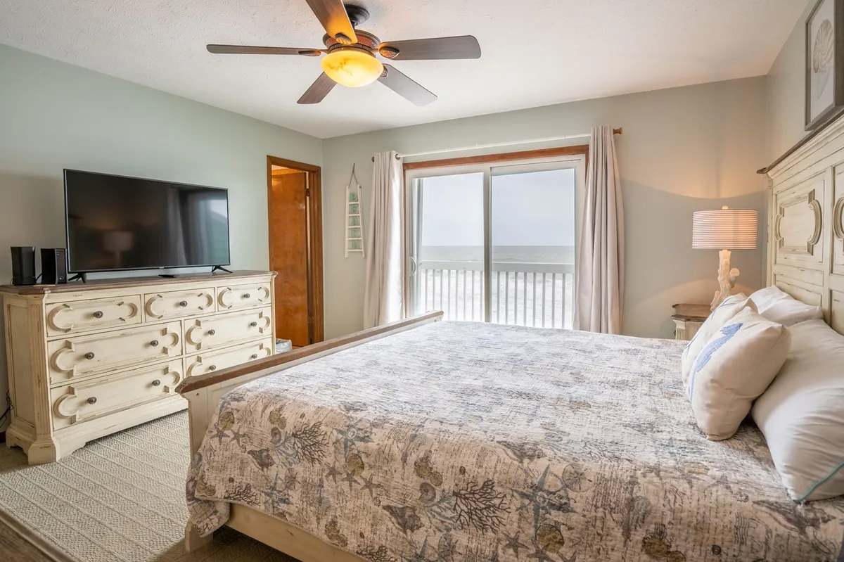 A serene bedroom featuring a bed with textured, patterned bedding positioned next to a large window framing expansive ocean views. A detailed dresser and soft, neutral decor enhance the tranquil atmosphere, creating a private retreat feel. The room is bathed in natural light, inviting restful relaxation and a connection to the ocean.