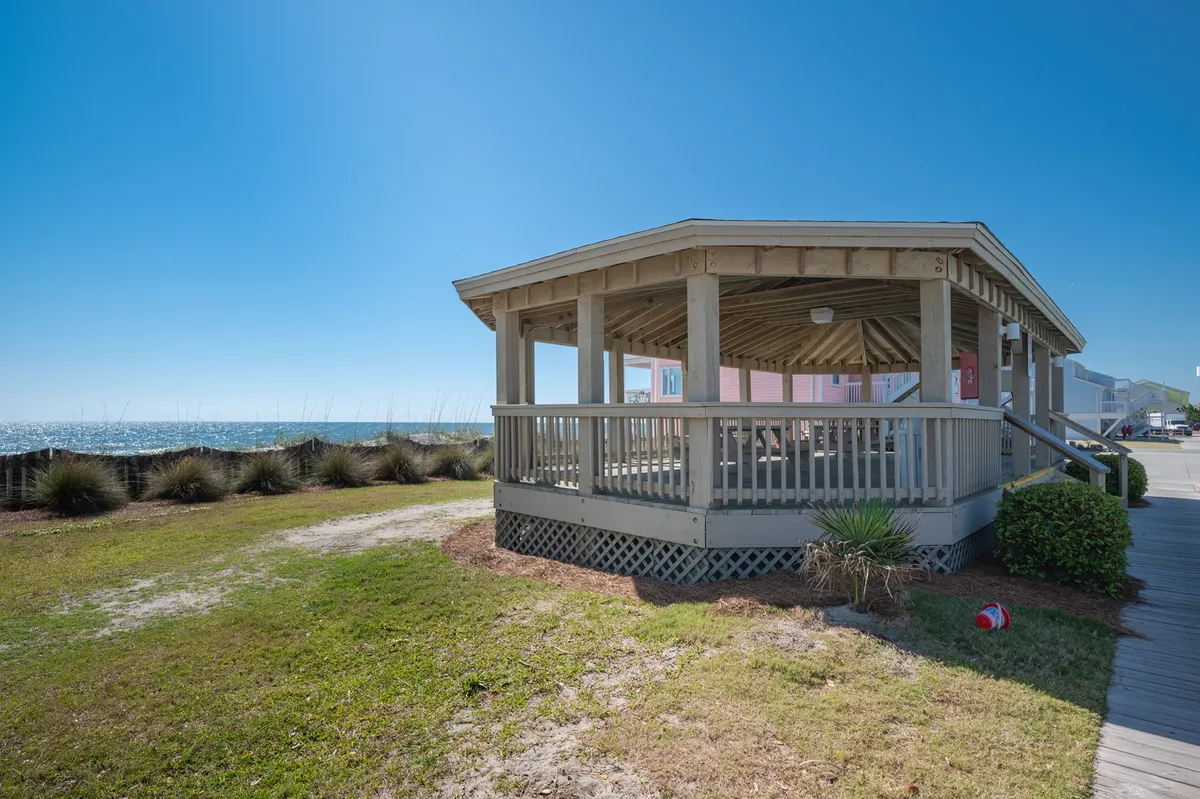 This image captures an octagonal wooden structure set on green grass, framed by lush palm plants and offering a spectacular ocean view. The expansive wooden deck is perfect for gatherings, dining, or simply soaking in the island scenery.