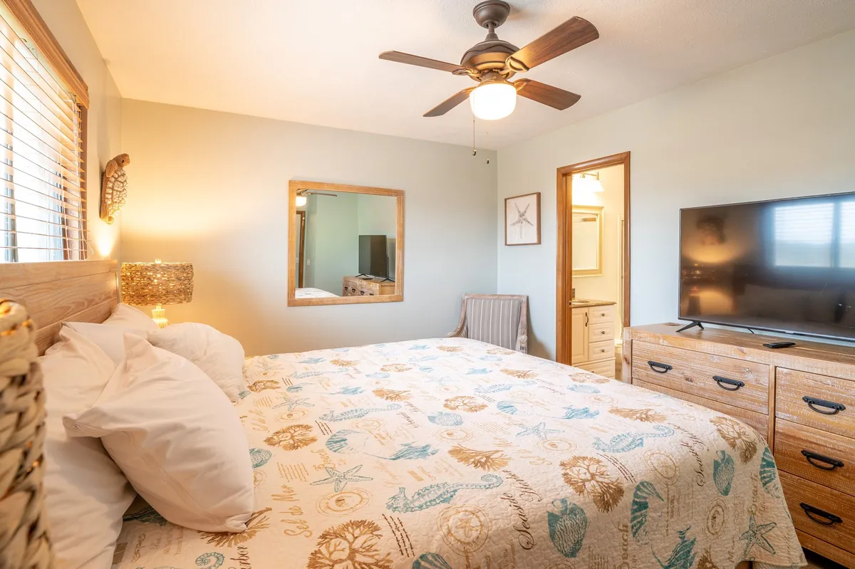 A guest bedroom featuring a plush bed adorned with decorative bedding, a TV on a wooden dresser, and framed artwork. The space feels inviting and tranquil, offering comfort and in-room entertainment, with a mirror reflecting natural light to create an open, relaxing ambiance.