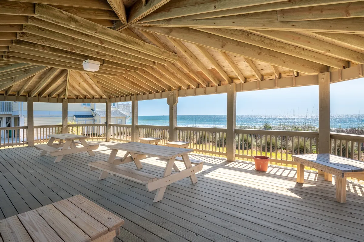 A relaxed, communal exterior space featuring wooden picnic tables under a covered structure with an open view of the ocean. The scene evokes togetherness and is ideal for family dinners, sunset gatherings, or casual meals by the beach.